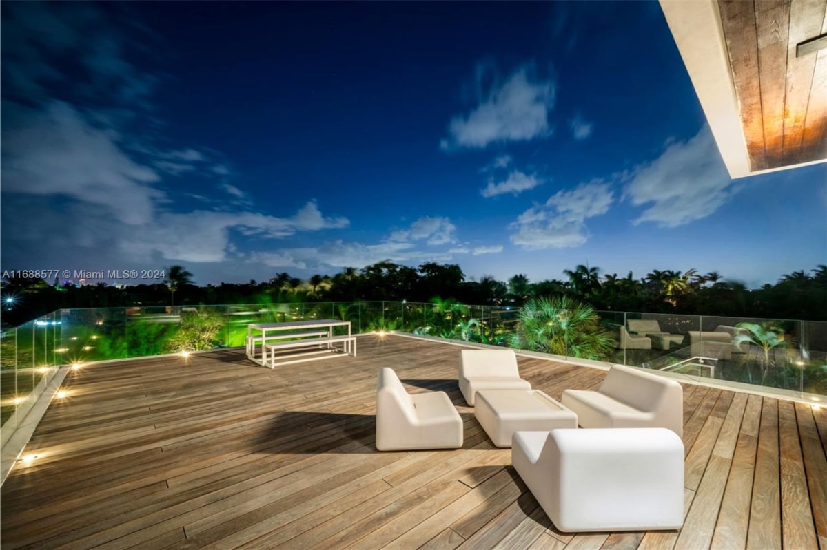 Nighttime view of a wooden deck with modern, white outdoor furniture. There's a white table and bench set, and a white sectional sofa arrangement.