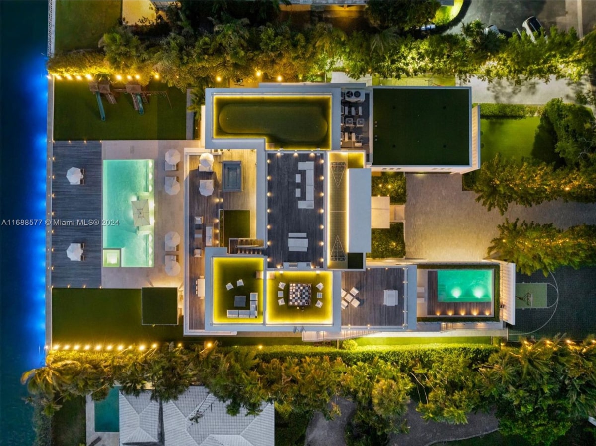 An overhead nighttime view of a building's rooftop. The rooftop appears to have multiple areas, including what seems to be a pool, lounge areas with seating, a game area, and other recreational spaces.