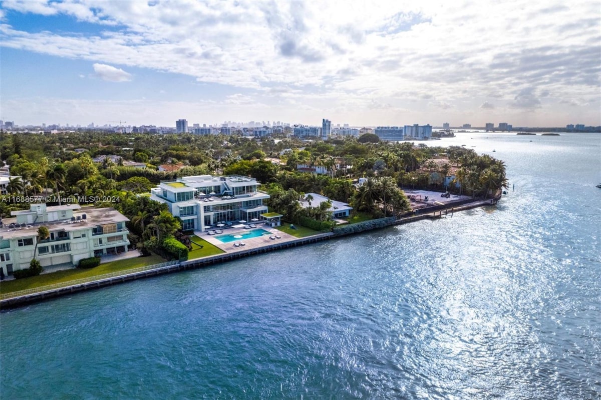 Aerial view of a large multi-storied, light-colored, and has a swimming pool. It's situated on a shoreline with lush greenery, and a cityscape is visible in the background.