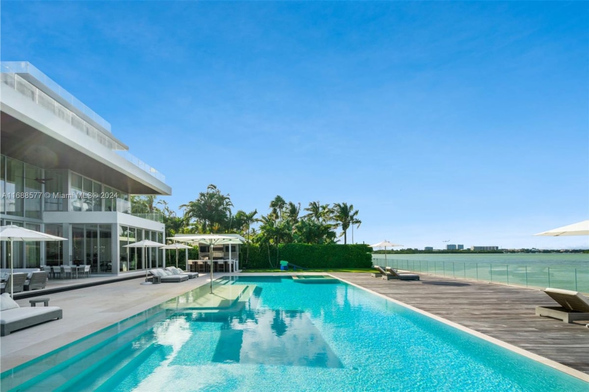 Rectangular swimming pool is the central feature, extending from a modern, white house towards a waterfront area with palm trees and a view of the water.