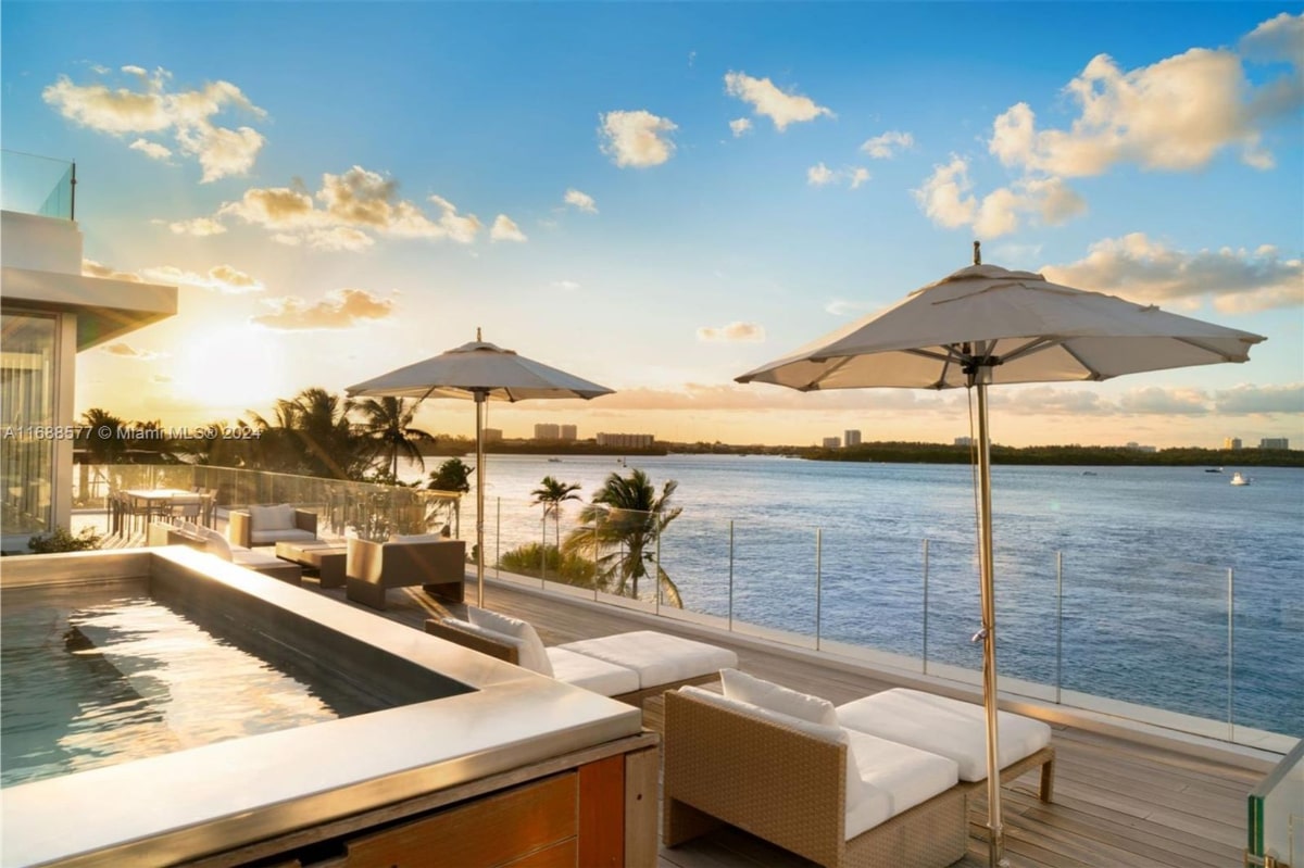 Outdoor patio area overlooking a body of water at sunset. There's a built-in pool or spa, comfortable seating, and large umbrellas providing shade.