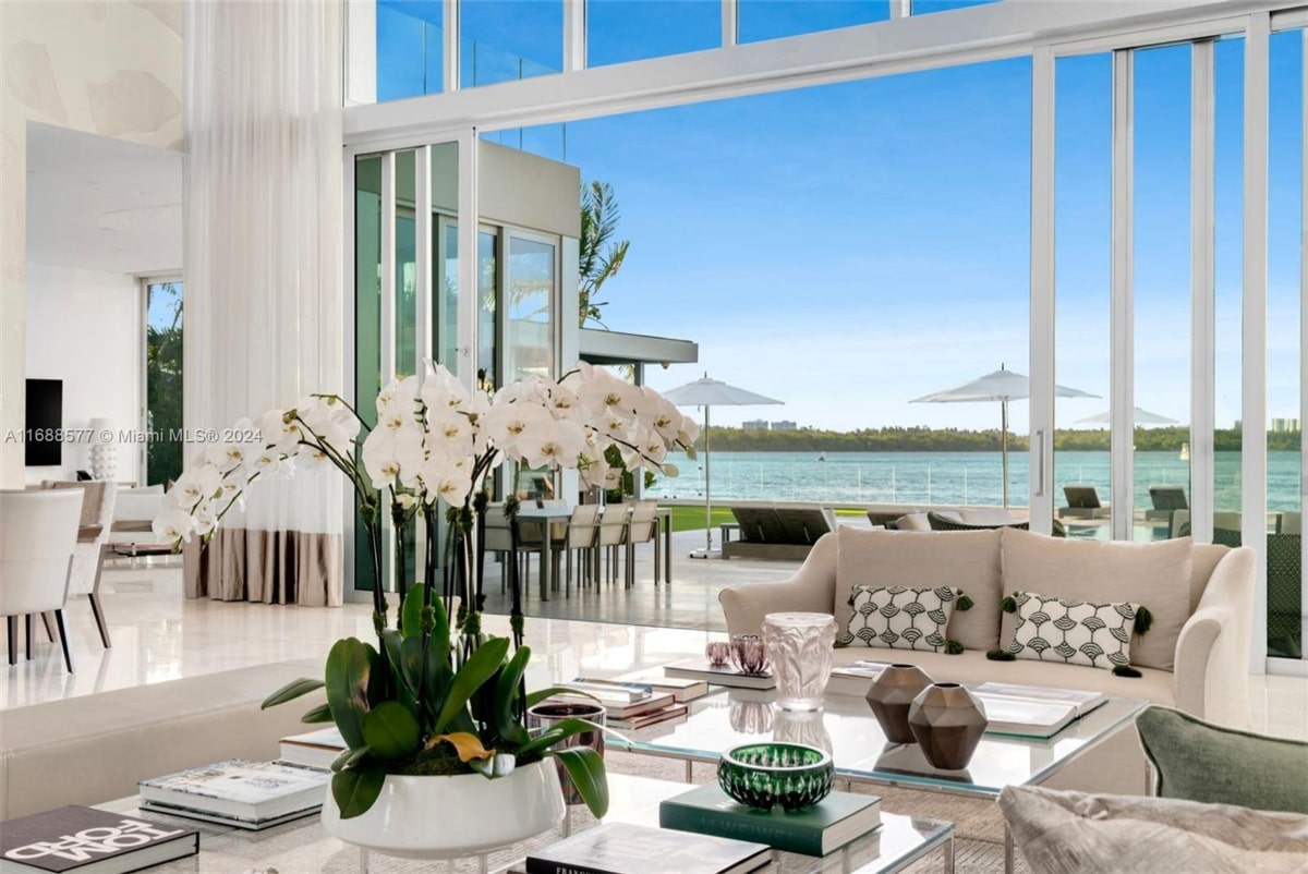 Interior mansion view of a waterfront. The room is decorated in a modern style with light-colored furniture and a large orchid plant as a centerpiece. Large windows offer an expansive view of the water and outdoor patio area.