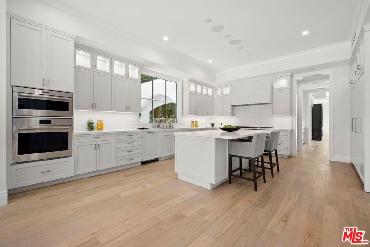 A modern kitchen with light gray cabinets, stainless steel appliances, and a large central island. The flooring is light wood.