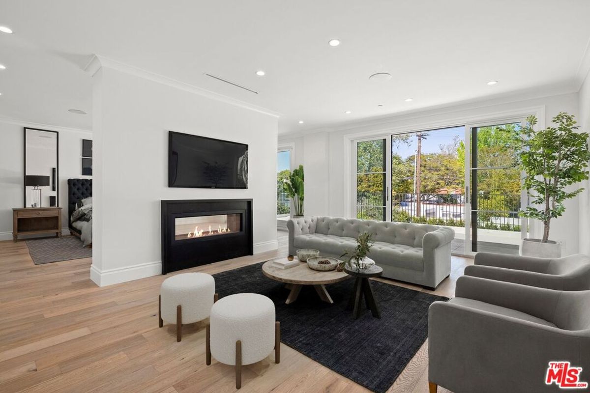 Living room with a large sofa, coffee table, and fireplace. The room has large windows and light wood floors. There is a glimpse of a bedroom in the background.