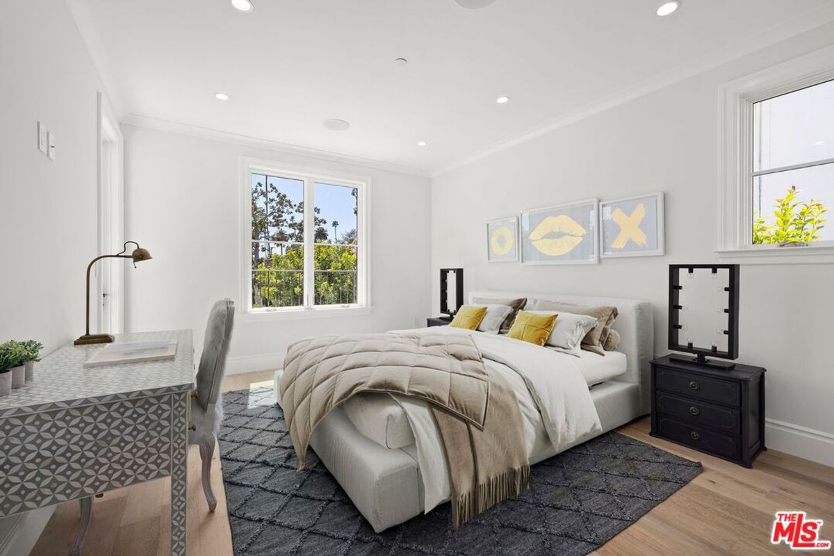 Bedroom with a large bed, a desk, and a window overlooking trees. The walls are white, and the floor is light wood.