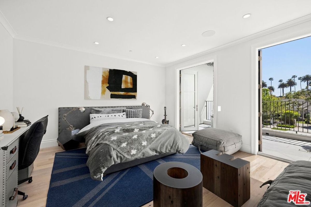 A bedroom with a bed, desk, and access to a balcony overlooking palm trees. The room has light wood floors and white walls.