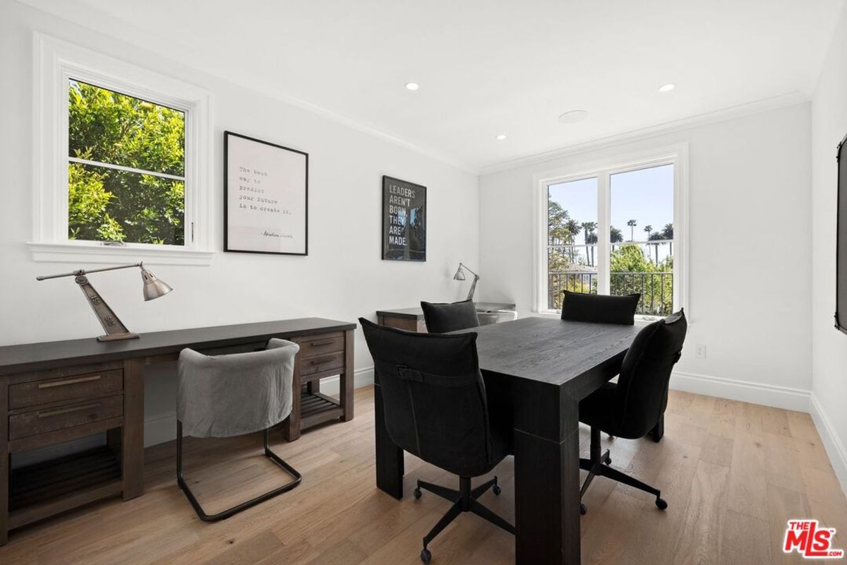 Home office with a large desk, a meeting table with chairs, and a window with a view of trees. The walls are white, and the floor is light wood.