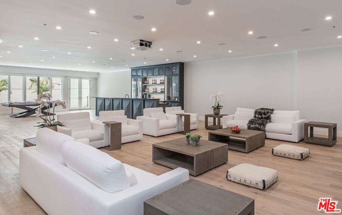 Large, open-plan game room with multiple white sofas, coffee tables, and a bar area. There are large windows and a pool table visible in the background.