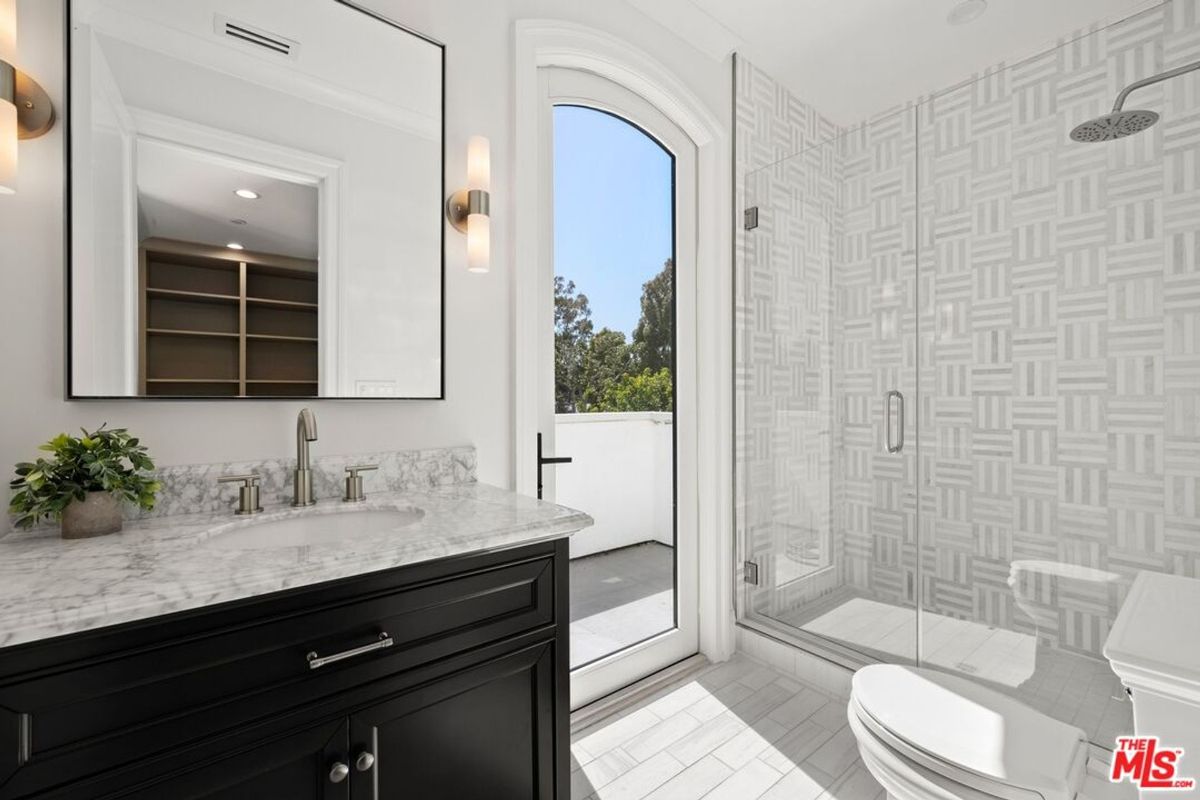 Bathroom with a vanity, a large mirror, and a walk-in shower with patterned tile. There is a door leading to an outdoor area.