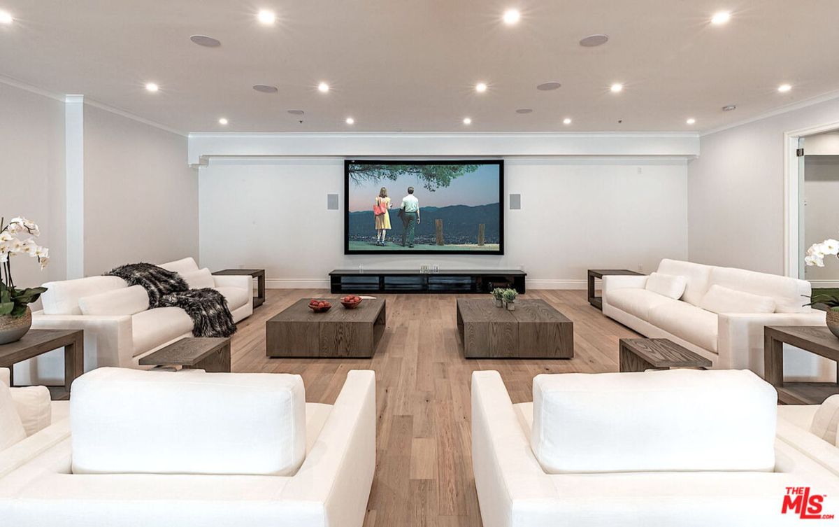 Home theater with white sofas, coffee tables, and a large projection screen showing a movie. The floor is light-colored wood.