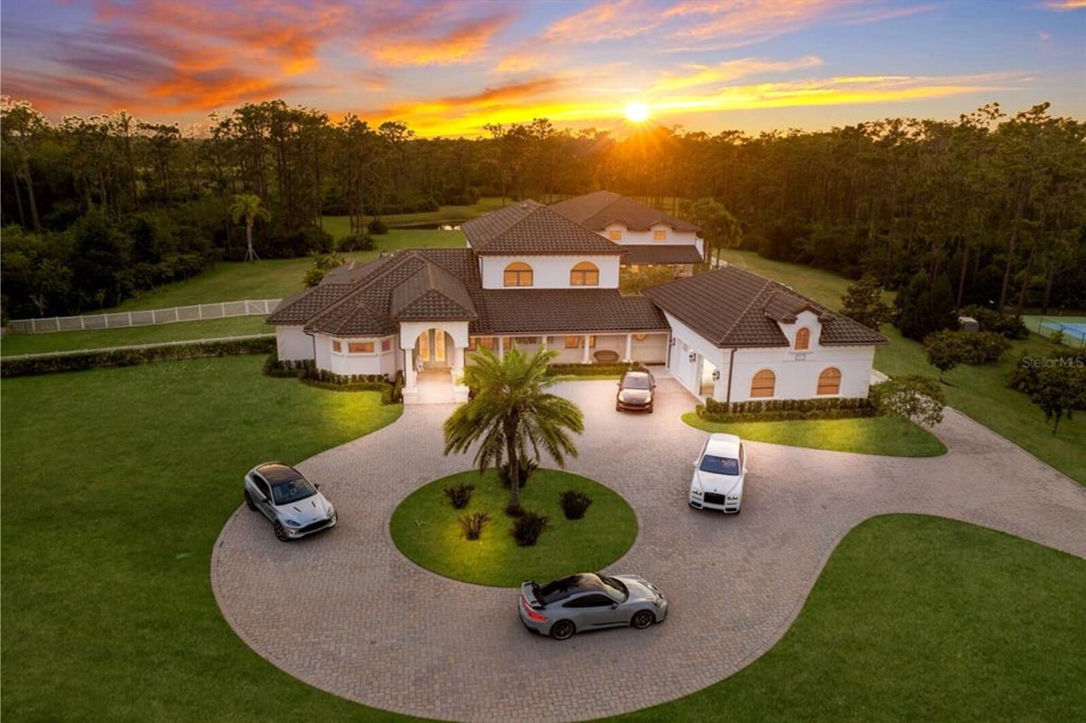 Aerial shot at sunset shows a large, luxurious white house with a brown tile roof, surrounded by a manicured lawn and several luxury cars parked in a circular driveway. A palm tree sits in the center of the circular driveway.