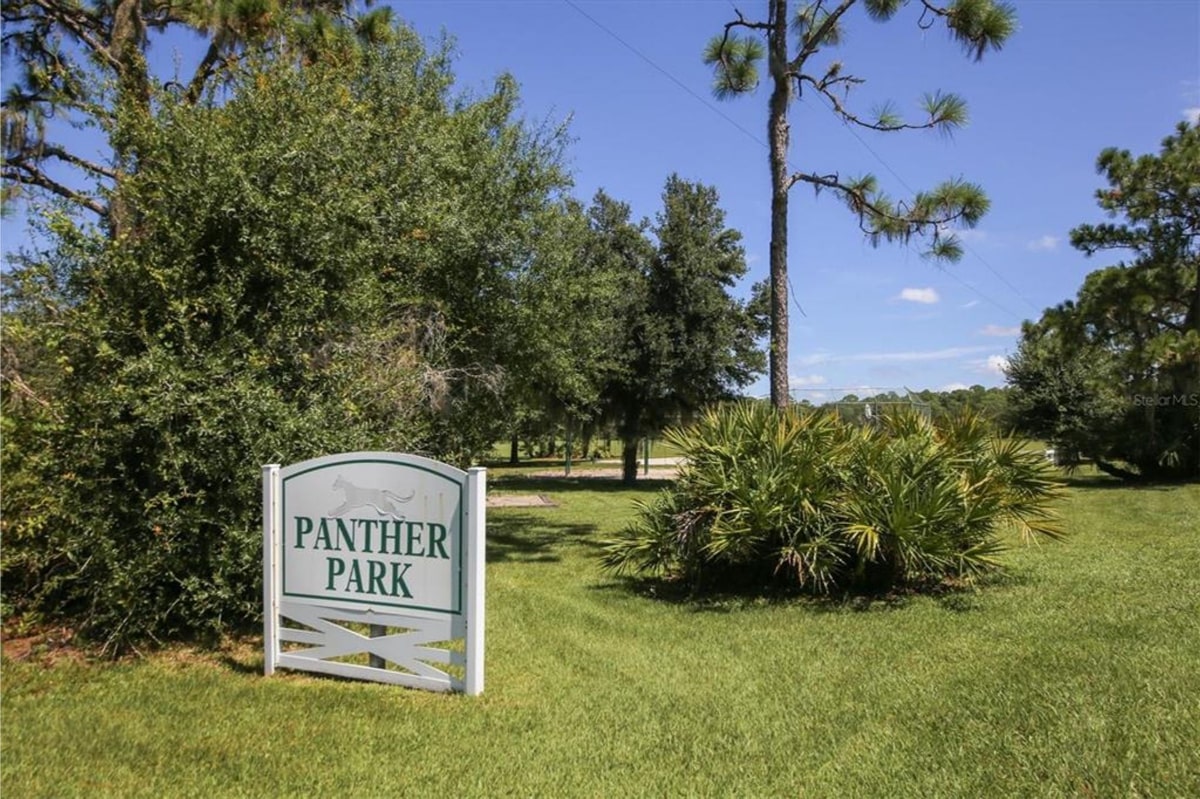 Sign for "Panther Park" in a grassy area with trees and some shrubs. The sign is white with black lettering. The setting appears to be a park or recreational area. The sky is clear and blue.