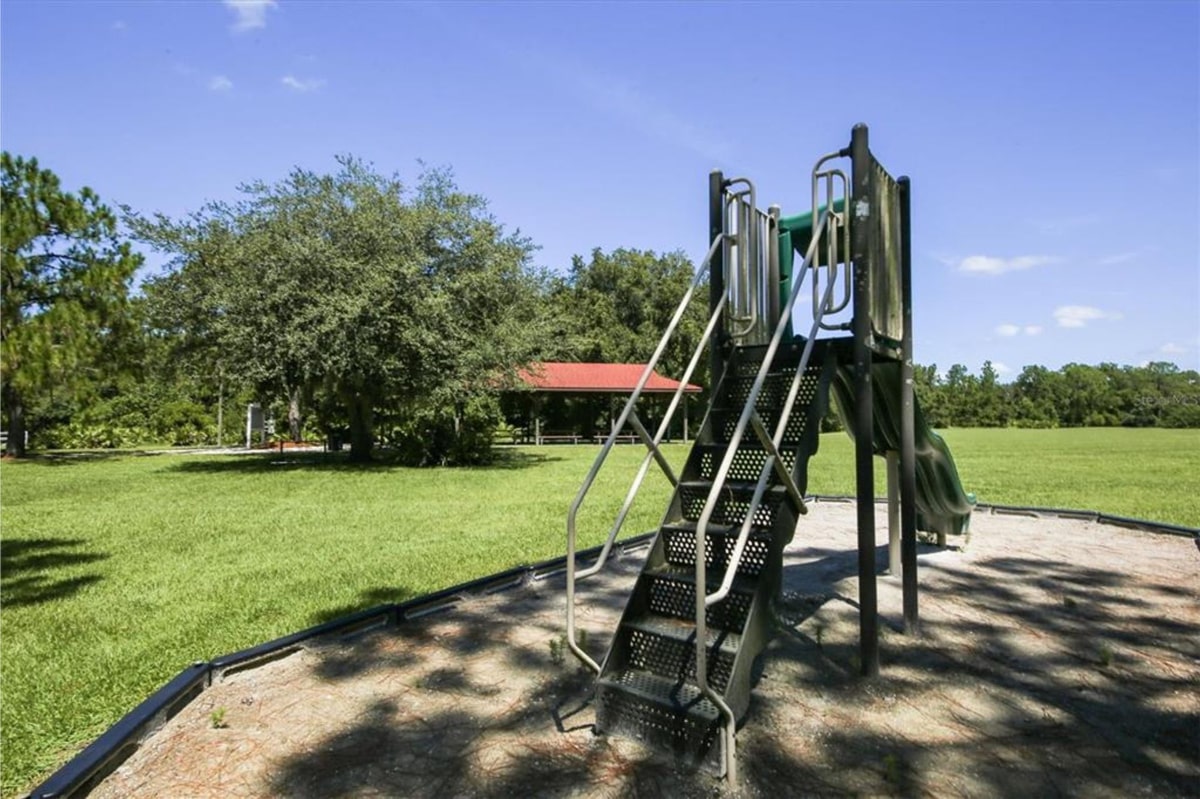 Playground slide in a park-like setting. The slide is dark-colored metal with stairs leading up to it. In the background, there's a grassy area, trees, and a small building that looks like a pavilion or shelter. The sky is clear and blue.