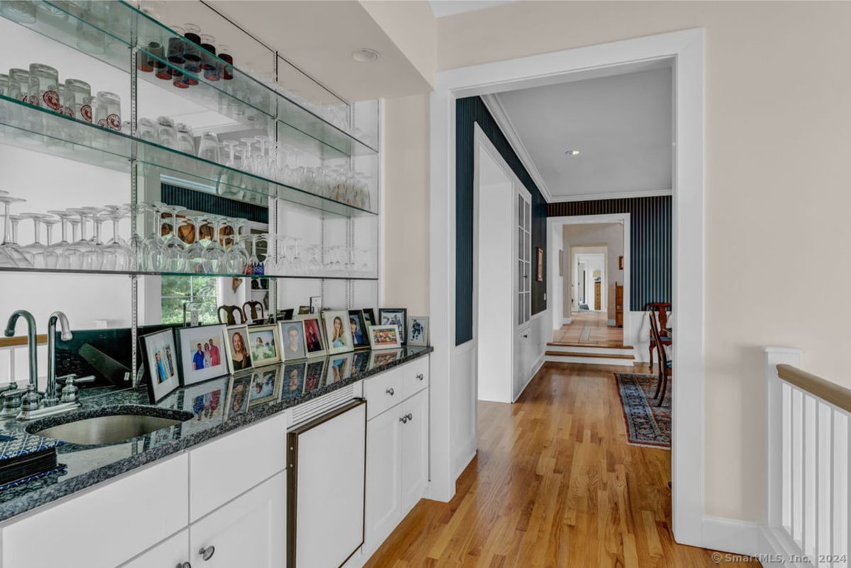 A long hallway with hardwood floors leads to a dining area, while a wet bar with glass shelving and family photos is situated to the side. The bar area features white cabinetry and a granite countertop.