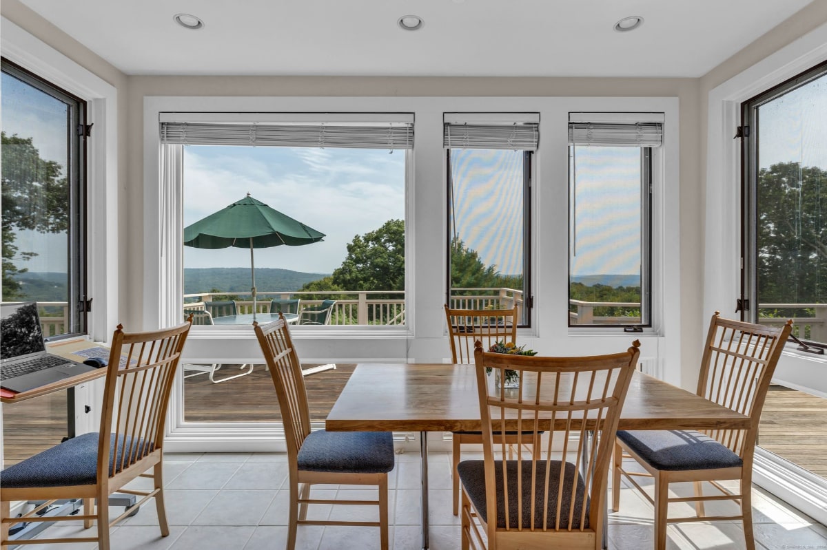 A sunroom with expansive windows offers stunning views of a deck and distant landscape. A wooden dining table with matching chairs is positioned to maximize the view.