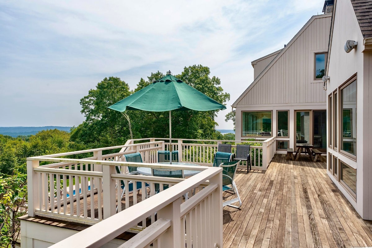 Deck overlooks a scenic vista, complete with a green umbrella shading a patio table and chairs. The deck is bordered by a white railing, and a house is visible in the background.