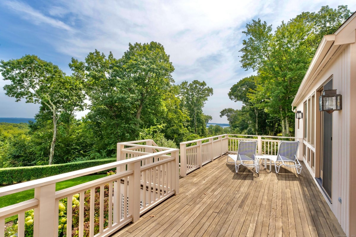 Large deck with white railings overlooks a tranquil seascape and lush greenery. Two lounge chairs are positioned to enjoy the expansive view.