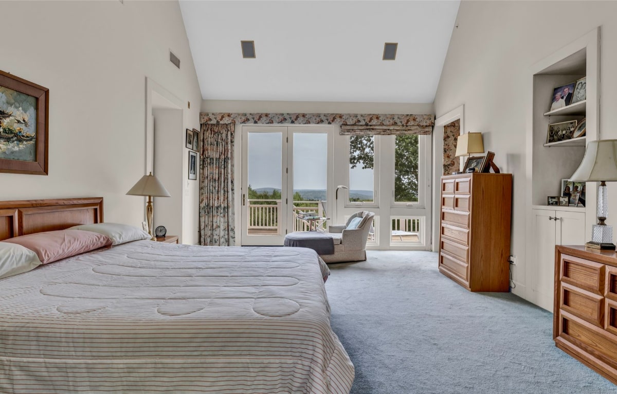 Master bedroom boasts a king-size bed with a striped comforter and access to a private balcony offering scenic views. The room features high ceilings, wood furnishings, and a calming color palette.