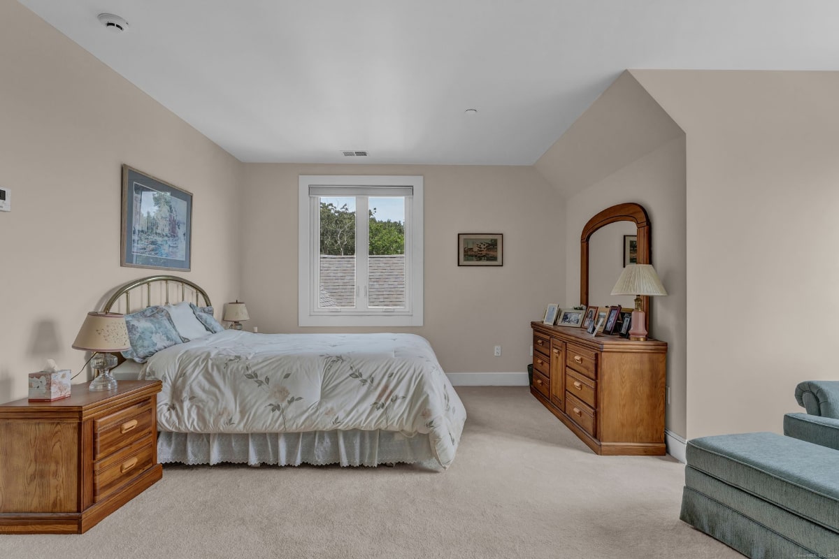 Queen-size bed with a light floral comforter, nightstands, and a dresser with a mirror. The room is spacious and has a neutral color scheme. A window provides natural light.