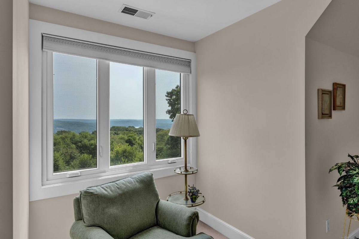 Tranquil seating area features a comfortable armchair positioned in front of a large window offering a picturesque view of a lake or ocean and surrounding landscape. A small side table with a lamp completes the scene.