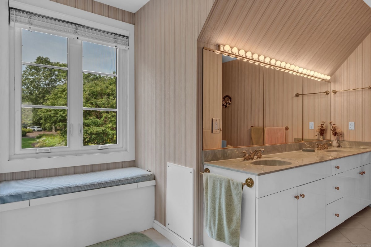 Bathroom with double vanity with a long mirror and a row of lights above. A window with a built-in window seat offers natural light and a view of the outdoors.