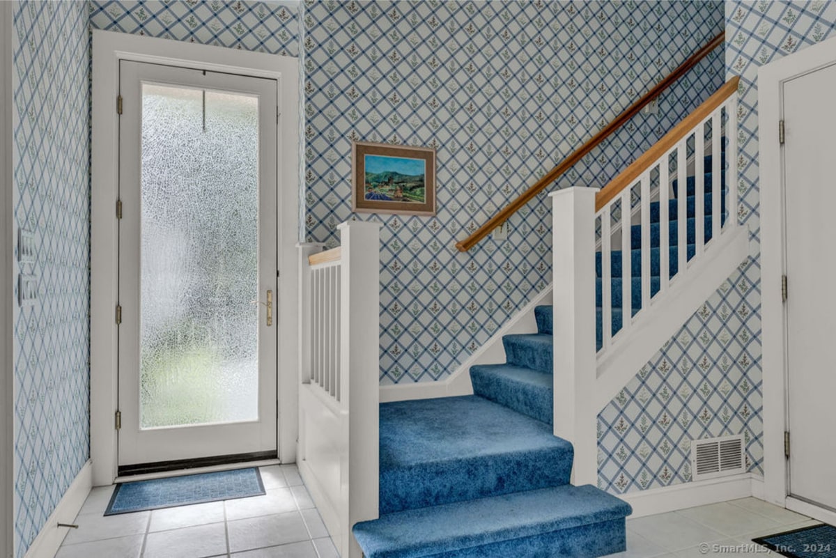 Entryway features a staircase with teal carpeting and a white railing. The walls are covered in patterned wallpaper, and a frosted glass door is visible to the left.