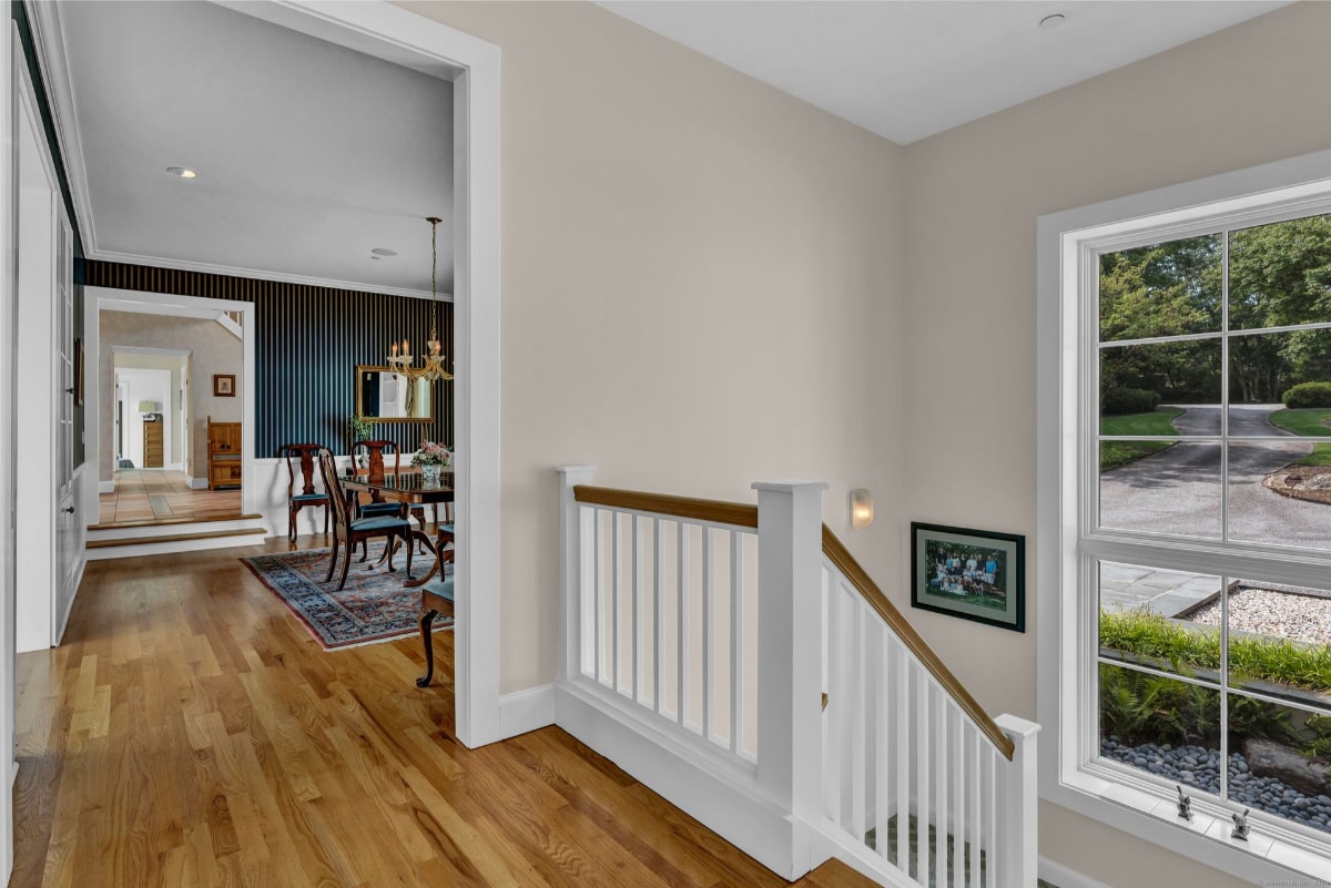 Bright interior with wooden flooring and a staircase with white railings. In the background, there's a dining area with a classic-style table and chairs, a chandelier, and dark accent walls.