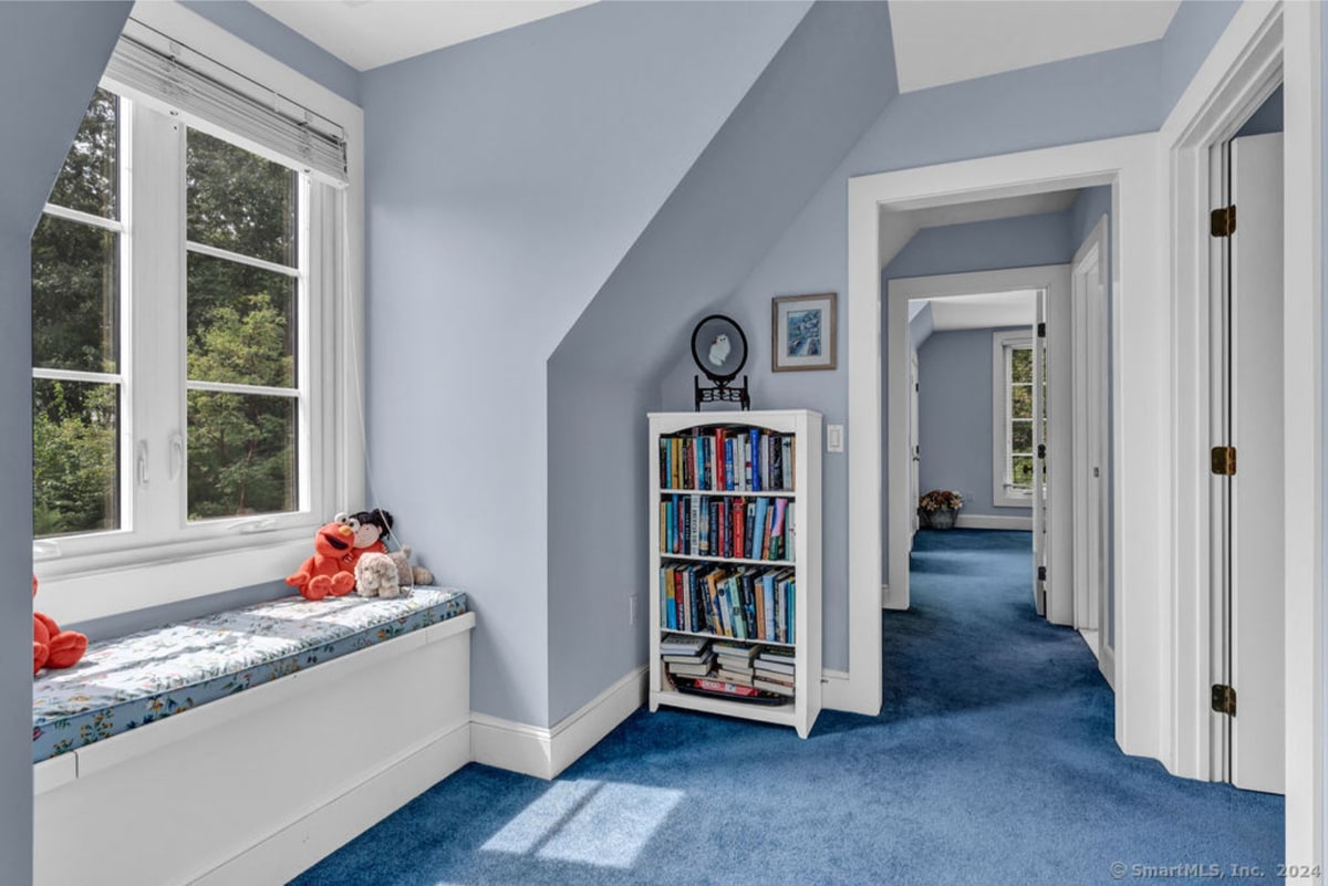 Cozy space with blue carpet and walls, featuring a window seat adorned with plush toys. A small bookshelf filled with books stands against the wall, leading into a hallway with additional rooms visible in the background.