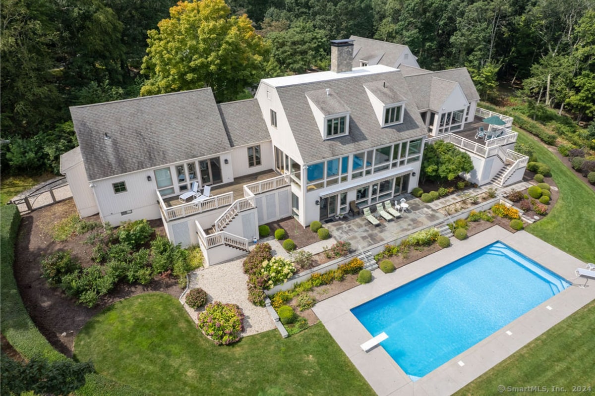 An aerial view of the white mansion that has multiple decks and stairs and is surrounded by landscaping.