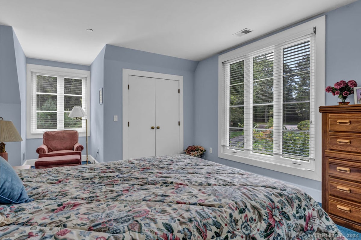 Serene bedroom with large windows that let in natural light, soft blue walls, and floral bedding. A cozy armchair sits near the corner, and a wooden dresser adds a warm touch to the space.