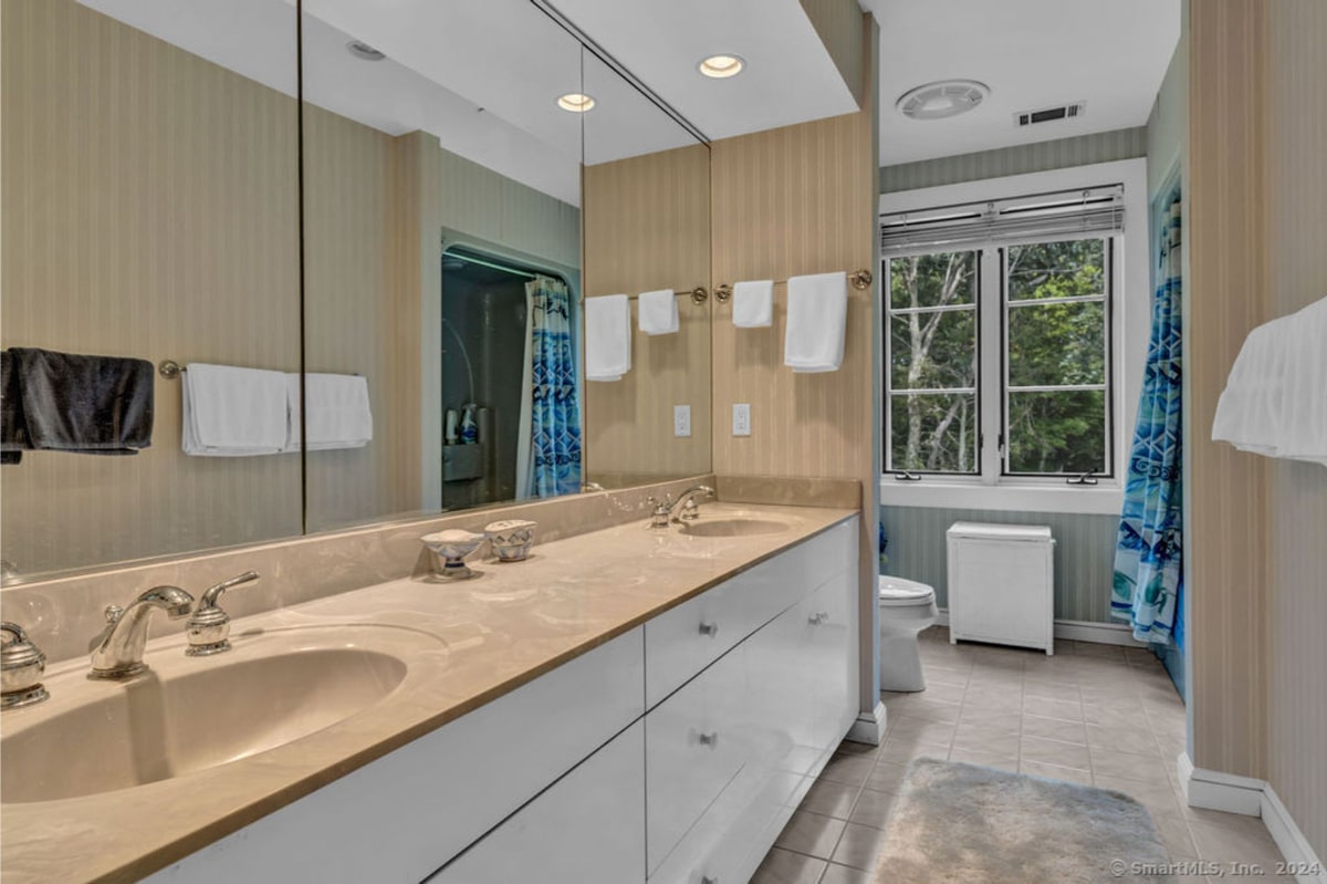 Bathroom with double sinks, white cabinets, large mirror, light and airy feel, with natural light streaming in from a window overlooking trees.