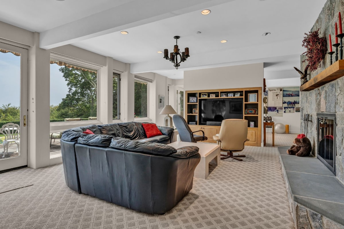 Family room with a large sectional sofa, built-in entertainment center, and a stone fireplace. Large windows offer a view of trees and greenery outside.