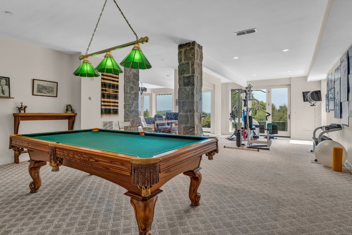 Game room with a pool table and a home gym. The room has large windows, stone columns, and a light-colored carpet.