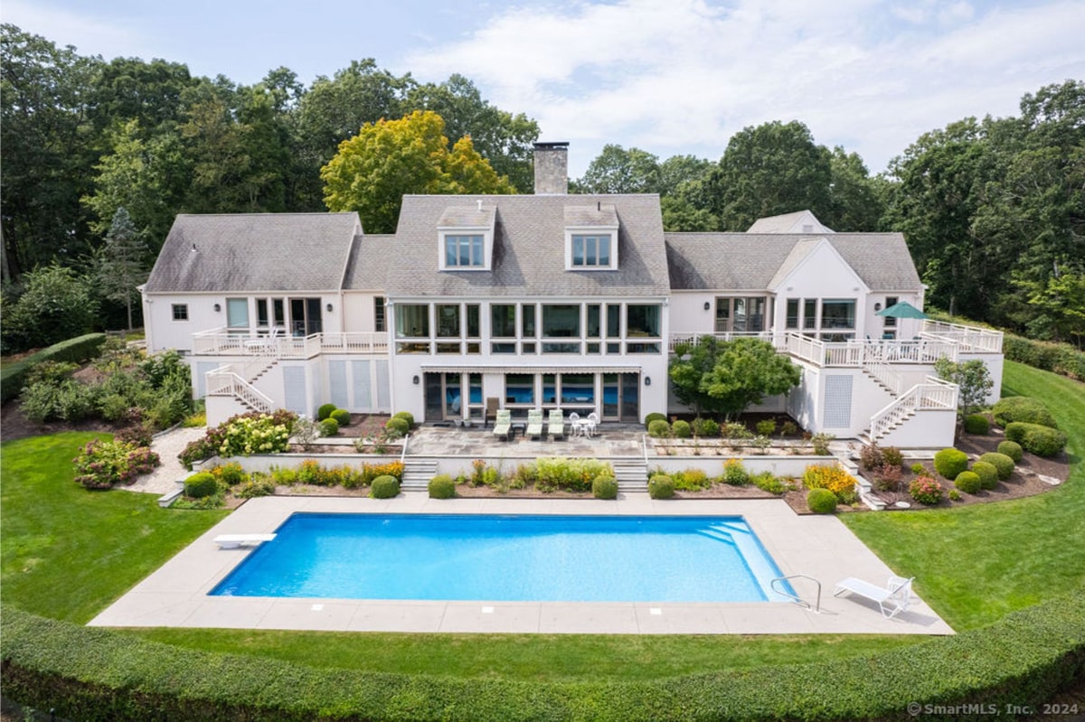 An aerial view shows a large, white two-story house with a swimming pool. The house has multiple levels, a large patio, and is surrounded by lush landscaping.