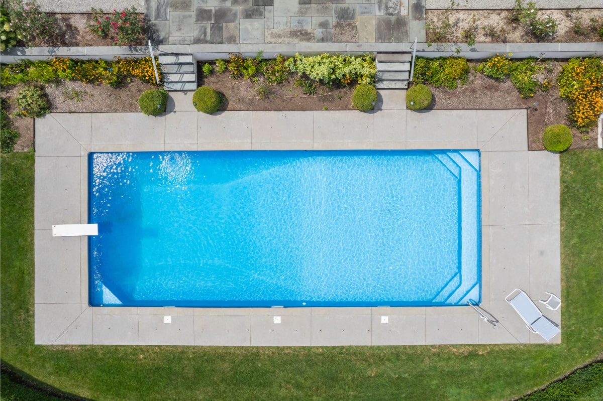 Bird's-eye view of a rectangular swimming pool. The pool is light blue, has a diving board, and is surrounded by a concrete deck and landscaping.