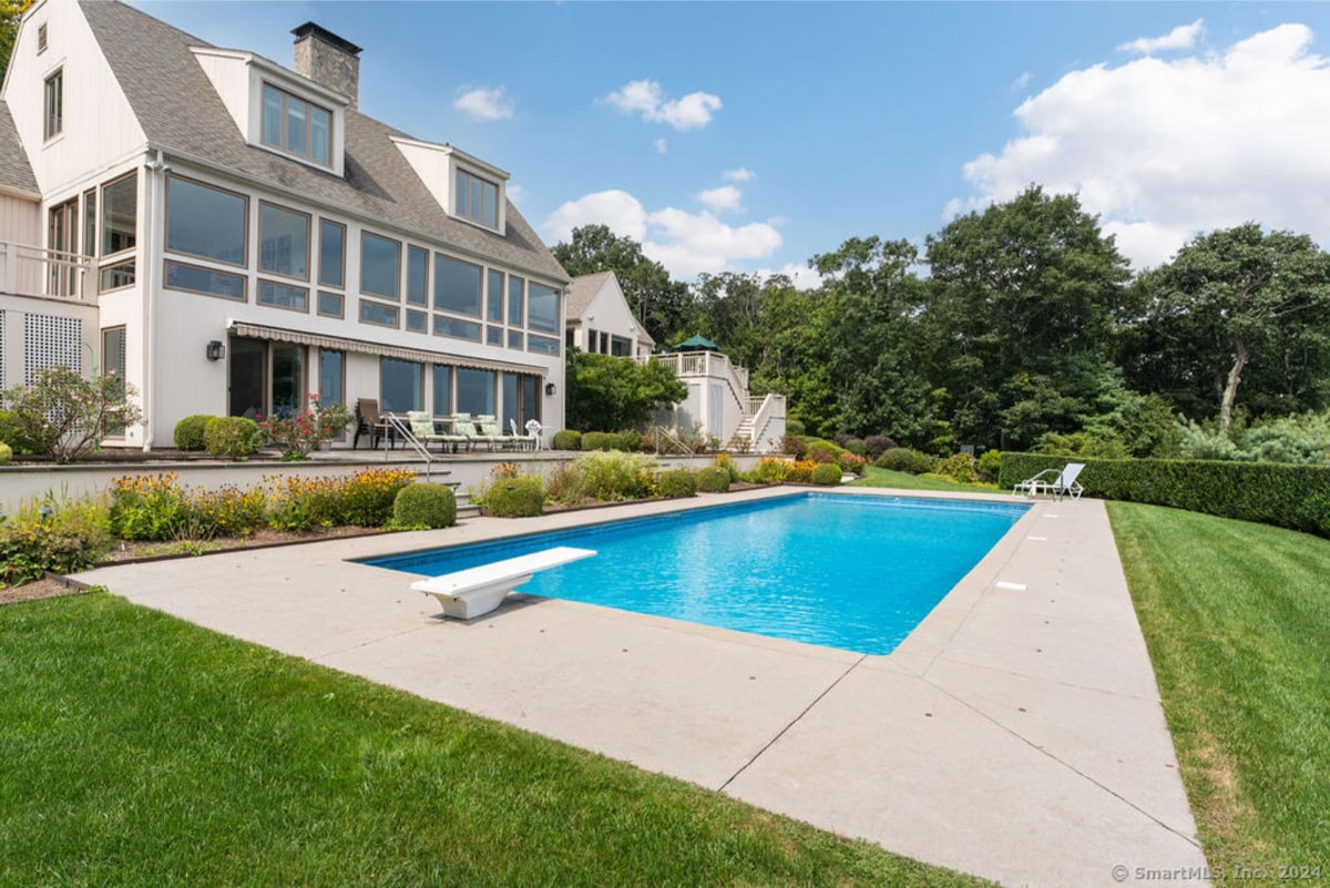 Light-colored mansion with a swimming pool in the backyard. The pool is rectangular with a diving board, and the property is surrounded by green lawns and trees.