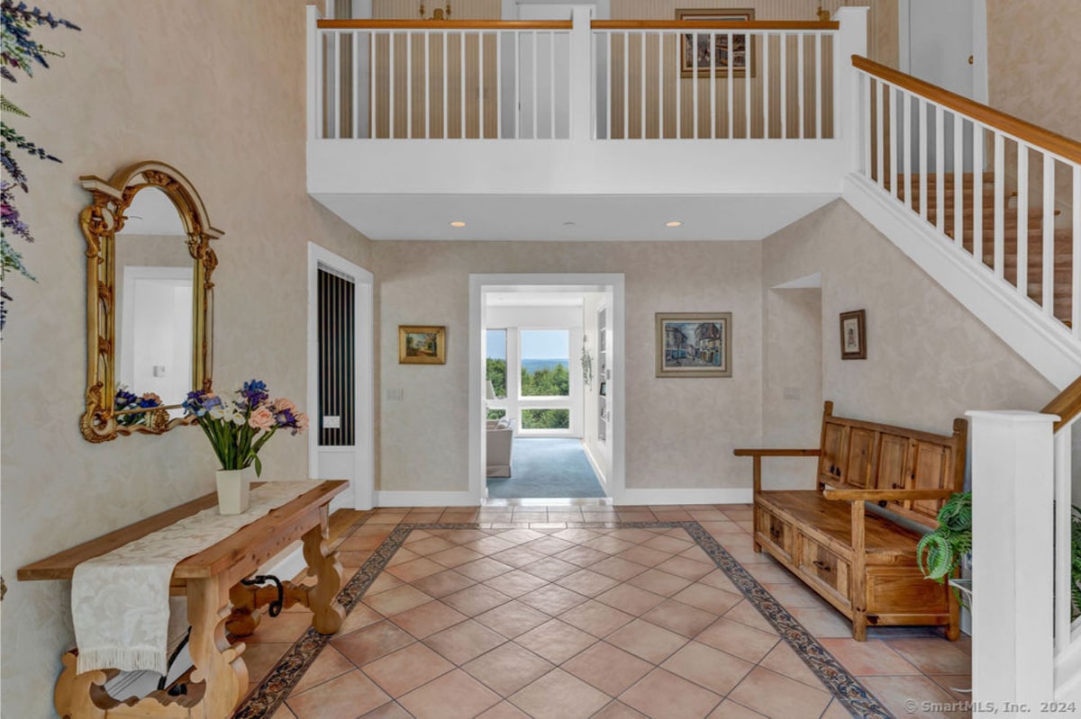 Bright, spacious foyer features a large mirror and wooden bench, with light beige tile flooring and a decorative border. A staircase ascends to an upper level, creating a sense of grandeur.