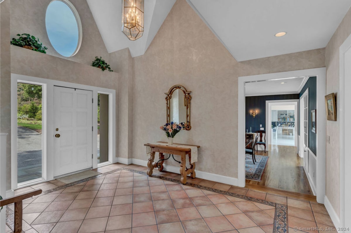 Home's entryway, showcasing high ceilings, a large chandelier, and an ornate mirror above a console table. Beyond the entryway, a view into a formal dining room with dark-colored walls is visible.