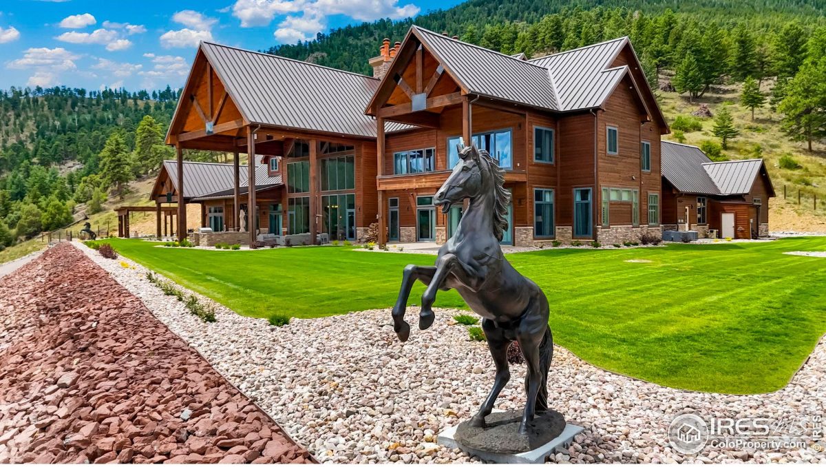 A rearing bronze horse statue stands prominently in front of a rustic-style home, set against a backdrop of mountains.