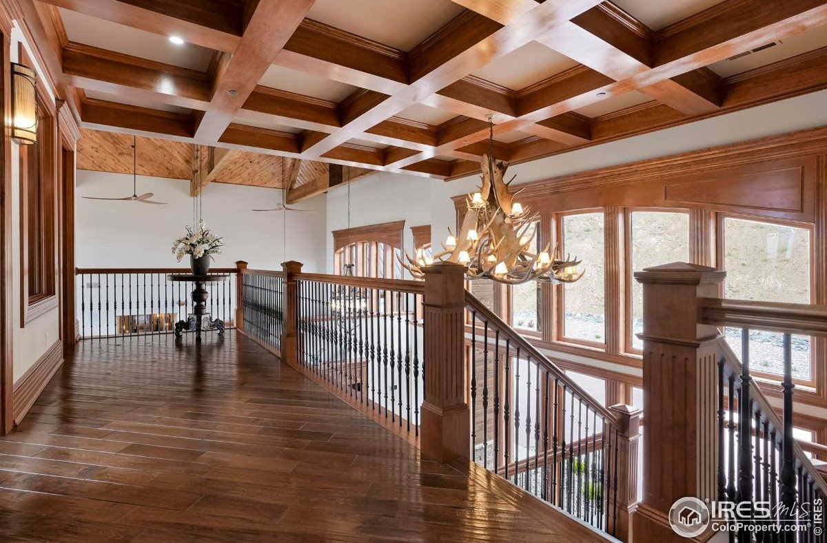 Upper-level hallway features a coffered ceiling, an antler chandelier, and a view overlooking a grand staircase.