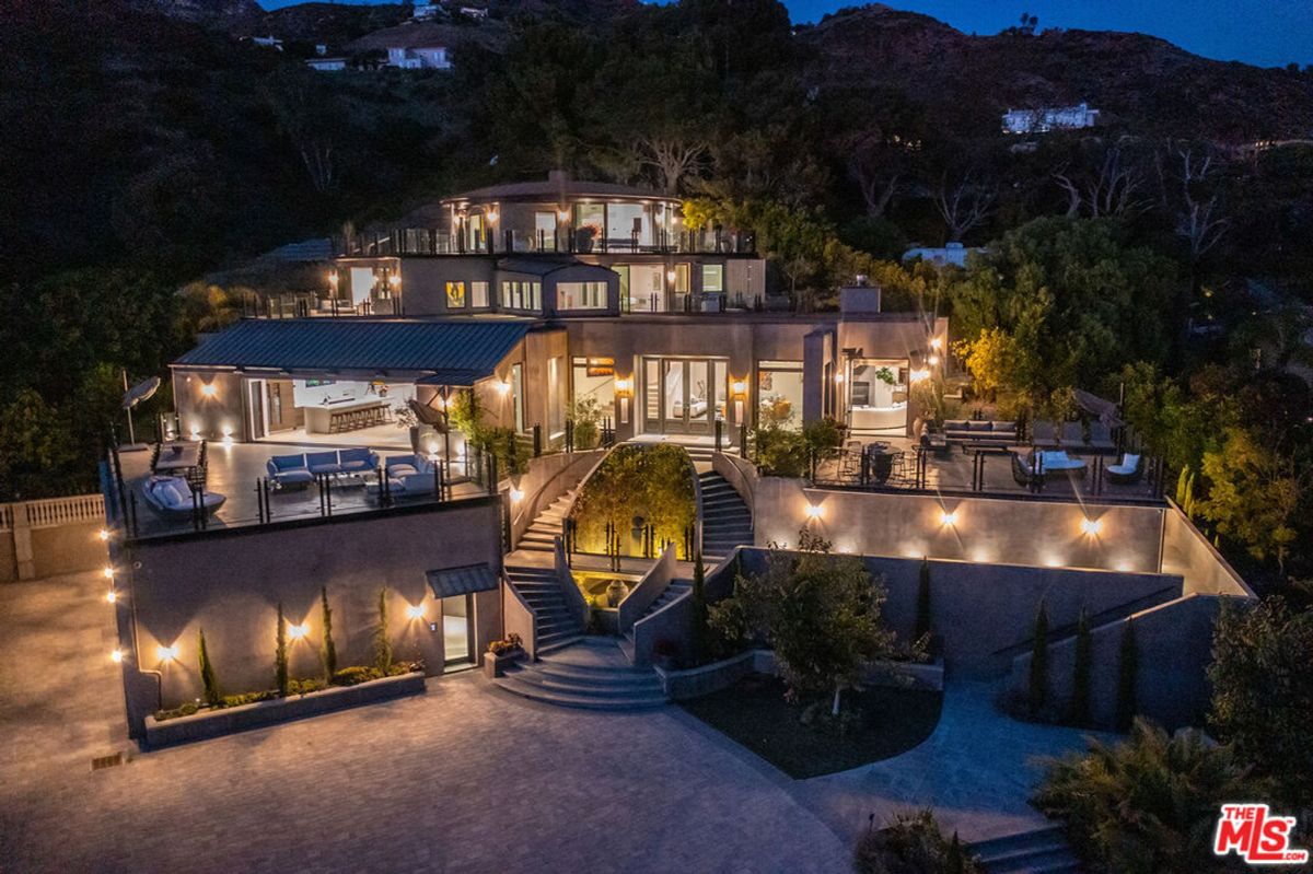 Multi-level modern mansion illuminated at night, nestled hillside, features expansive decks and dramatic staircase.