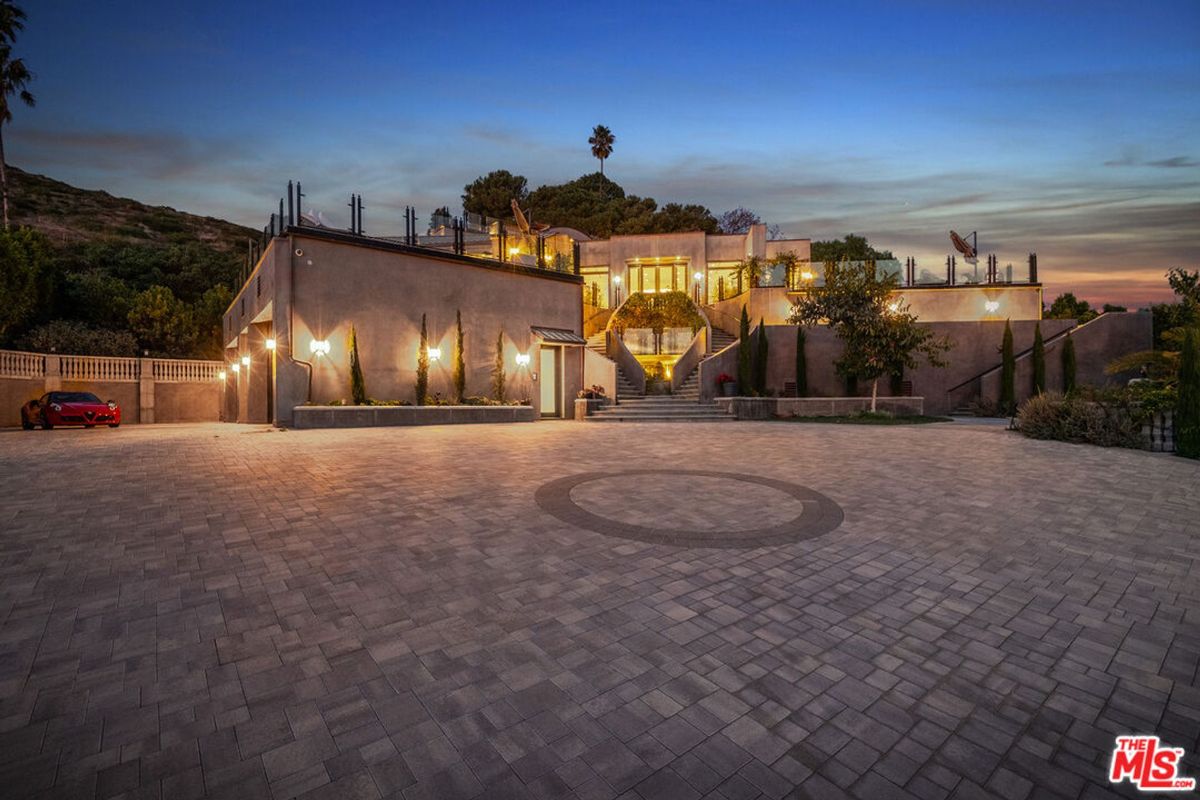 Expansive estate, dusk lighting, showcases grand architecture, expansive paved motor court, hillside setting.