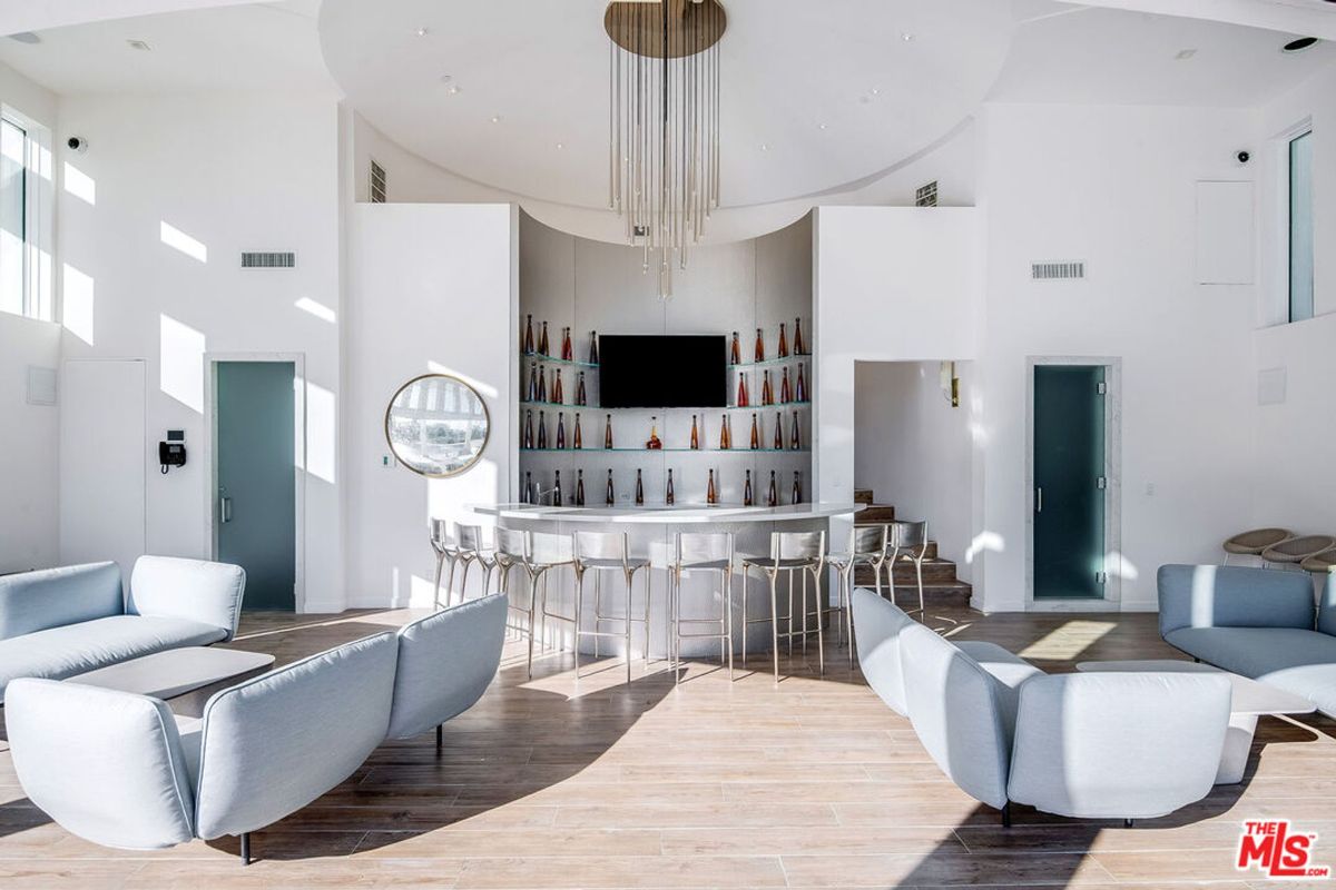 Modern bar area with a high ceiling, light-colored walls, and plenty of seating. A large chandelier is a focal point.