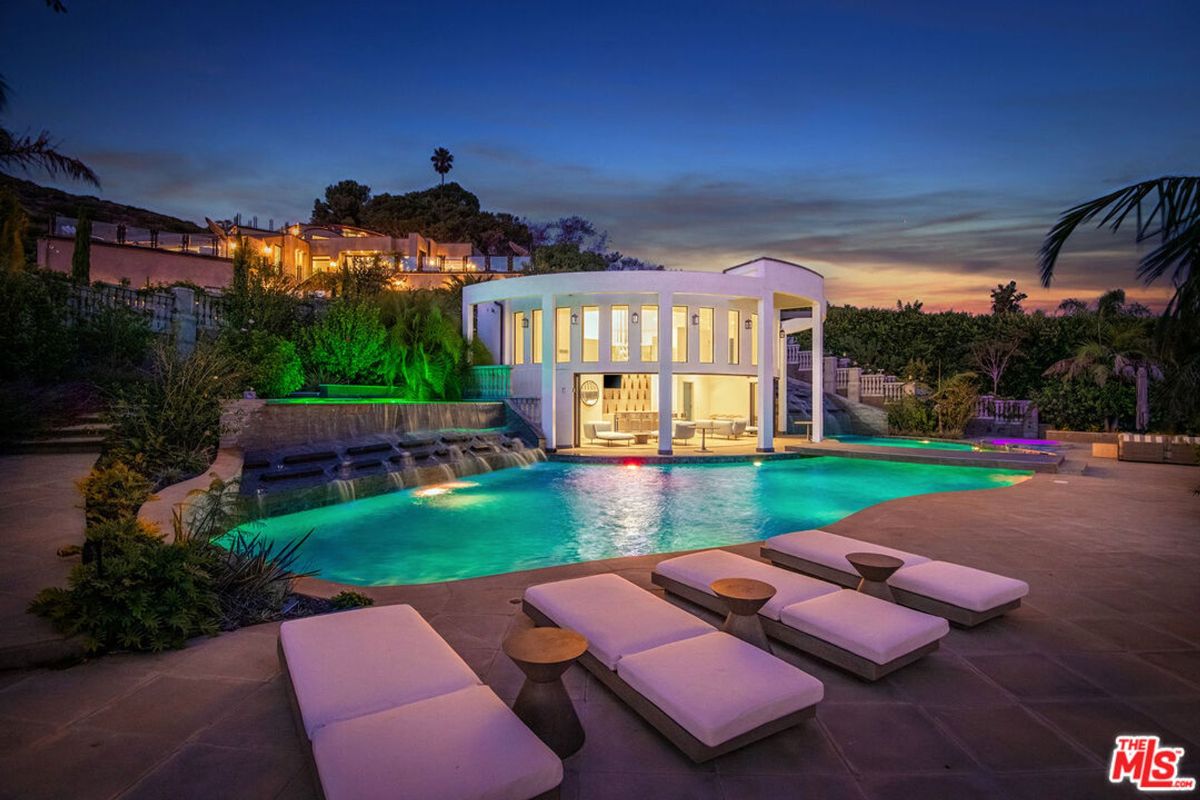 Nighttime view of a swimming pool and a modern house with lights on in the background. Lounge chairs are positioned around the pool.