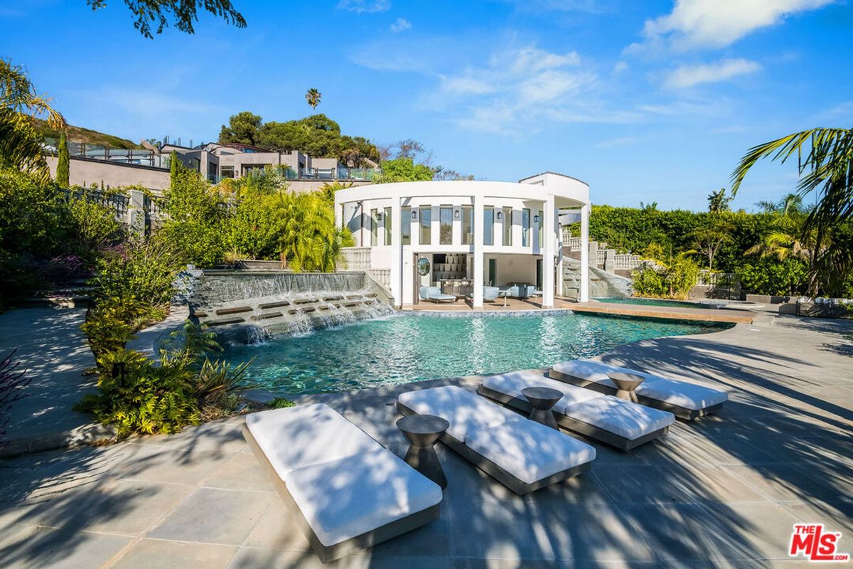 Luxury pool area with a waterfall feature, lounge chairs, and a modern pool house. The pool is surrounded by lush landscaping.