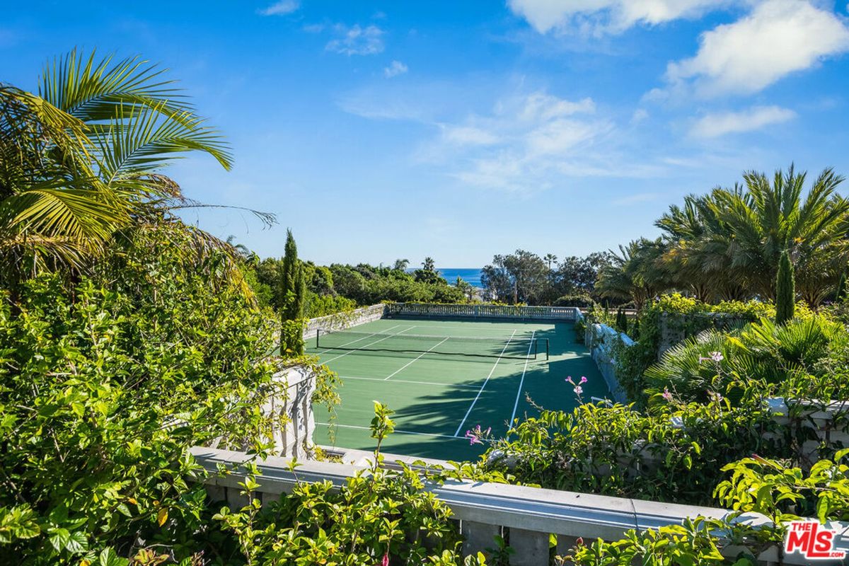 Elevated view of a tennis court with ocean views in the background. Lush greenery surrounds the court.