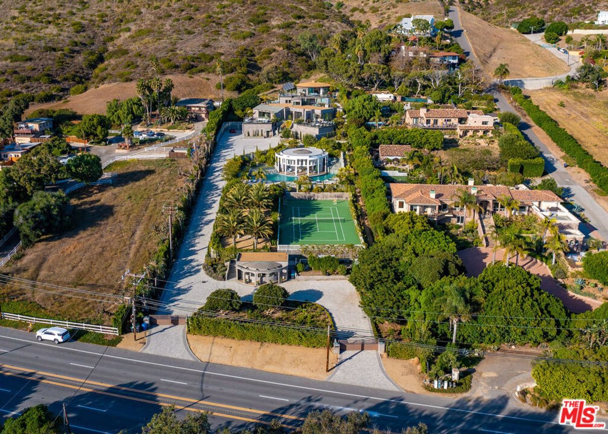 Aerial view of a luxury residential area with multiple houses, palm trees, a tennis court, and a swimming pool. The properties are situated on a hillside.