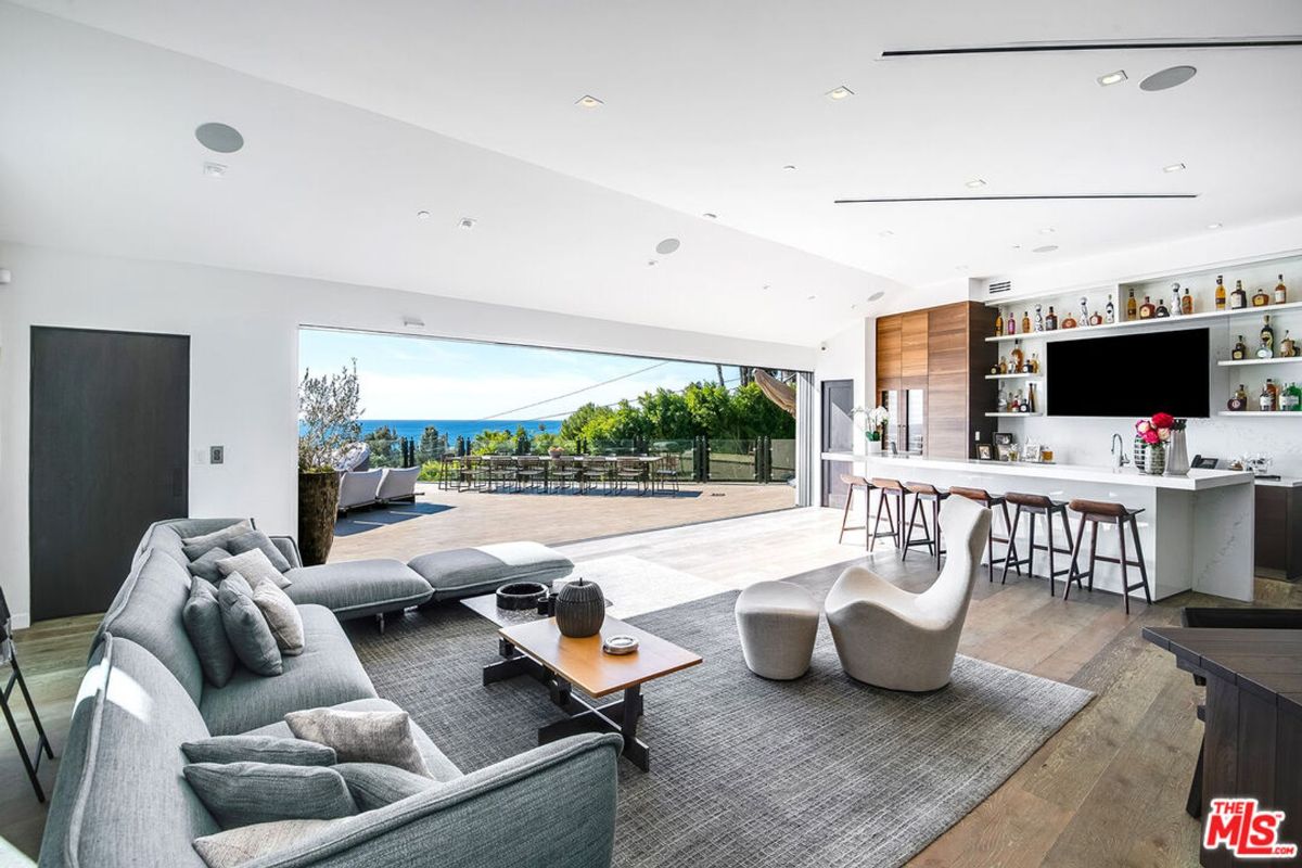 Expansive great room, floor-to-ceiling windows showcasing ocean views, features modern sectional sofa, wet bar, and hardwood floors.