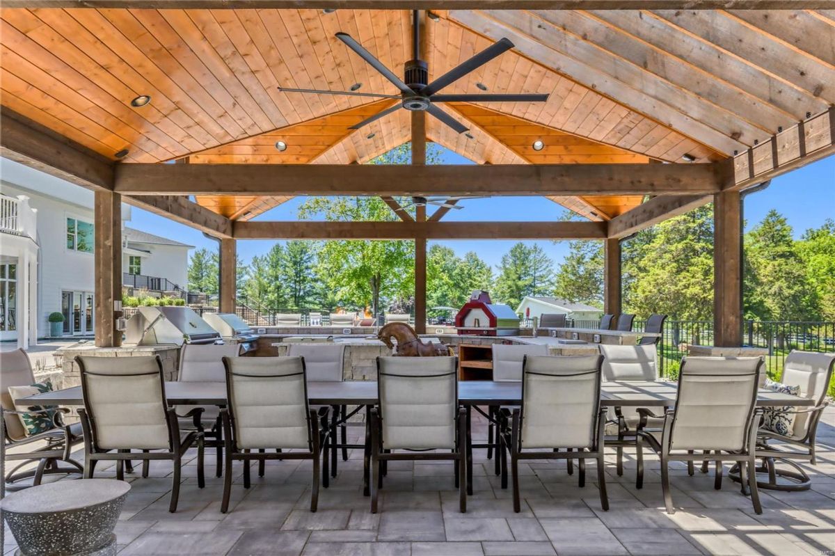 A large outdoor dining area is situated under a covered pavilion with a wood-beamed ceiling and a ceiling fan. 