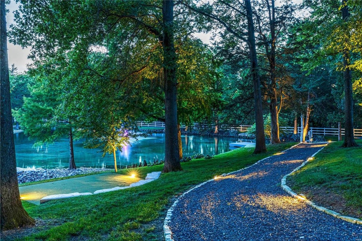 A gently curving pathway, lit by soft lights, winds through a wooded area beside a tranquil pond.